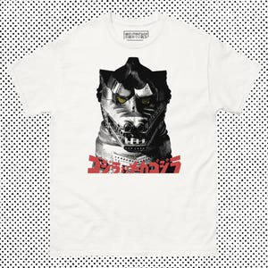 Puede incluir: Camiseta blanca con un gráfico en negro y plata de una cabeza de monstruo con ojos amarillos y texto japonés en rojo. La camiseta tiene cuello redondo y mangas cortas.