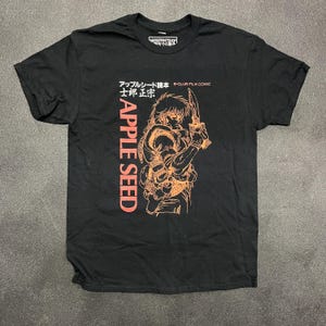 May include: Black t-shirt with an orange graphic of a character from the anime "Apple Seed". The text "Apple Seed" is written vertically in orange on the left side of the graphic. The text "B-CLUB FILM COMIC" is written in orange at the top of the graphic. The text "アップルシード  士郎正宗" is written in orange above the graphic.