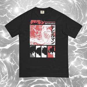 May include: A black short-sleeve t-shirt featuring a graphic print with the text "DEVILMAN" and Japanese characters. The design includes a large red and white illustration of a creature and four smaller panels showing character profiles.