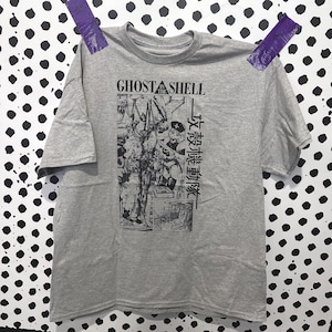 Könnte beinhalten: Ein graues, kurzärmeliges T-Shirt mit einem schwarzen Grafikdruck auf der Brust. Das Design enthält den Text GHOST IN THE SHELL und japanische Schriftzeichen neben einer detaillierten Illustration einer Figur in einer mechanischen Umgebung.