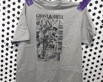 May include: A heather gray short-sleeve t-shirt featuring a black graphic print on the chest. The design includes the text GHOST IN THE SHELL and Japanese characters alongside a detailed illustration of a character in a mechanical setting.