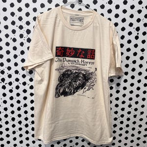 Könnte beinhalten: Cremefarbenes T-Shirt mit dem Text "The Dunwich Horror" von H.P. Lovecraft und japanischem Text darüber. Die Grafik ist eine Schwarz-Weiß-Illustration einer Kreatur. Das Shirt hat kurze Ärmel und einen Rundhalsausschnitt.
