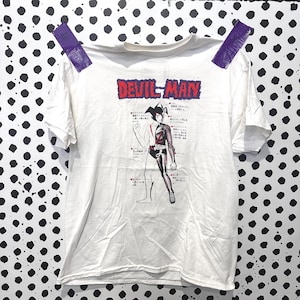 May include: A white short-sleeve t-shirt featuring a graphic of the character Devilman. The design includes the red text DEVIL-MAN at the top and a detailed anatomical diagram of the character with Japanese text labels pointing to various body parts.