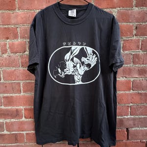 Może przedstawiać: Czarny t-shirt z krótkim rękawem z białym nadrukiem graficznym przedstawiającym postać w dynamicznej pozie wewnątrz owalnej ramki. Nad owalem znajduje się japoński napis "デビルマン" wydrukowany na biało. Koszulka posiada okrągły dekolt.