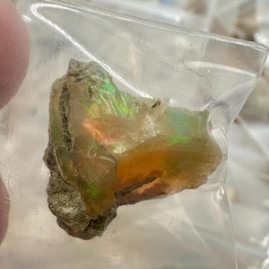 May include: A rough, iridescent opal gemstone fragment, encased in a clear plastic bag. The opal displays a spectrum of colors, including green, orange, and yellow, with a textured, earthy matrix. The gemstone is a natural, uncut specimen.