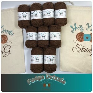 May include: Eight brown balls of yarn in a white drawstring bag with the text "My Kinda String" in brown and orange. The bag is on a teal surface with the text "Fudge Drizzle" in white.