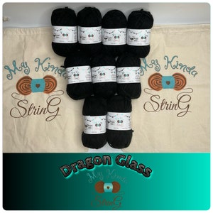 Puede incluir: Ocho ovillos de hilo negro en una bolsa de cordón blanco con el texto "My Kinda String" y un lazo de hilo marrón y verde azulado. La bolsa está sobre un fondo verde azulado con el texto "Dragon Glass" y el mismo logotipo que la bolsa.
