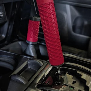 May include: A pair of red cylindrical gear shift and handbrake handles featuring a textured diamond waffle pattern. The handles have black caps at the top and bottom and are installed on the center console of a vehicle with a black gear shift gate.