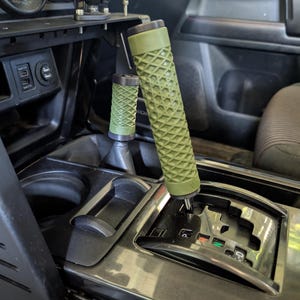 May include: Interior shot of a vehicle's console featuring a gear shift and a smaller shift knob, both with olive green diamond-patterned grips. The console is black with silver accents, and the image is well-lit.