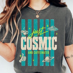 May include: Dark gray t-shirt with the text "just COSMIC and caffeinated" in a retro design. The graphic includes a coffee cup, a UFO, a thumbs-up, and a planet with rings. The design is set against teal vertical stripes.