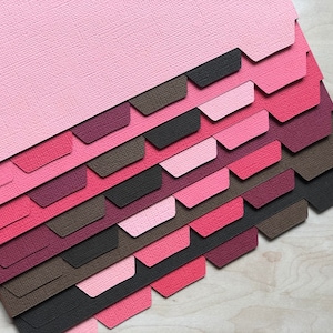 May include: A stack of colorful paper dividers in shades of pink, red, and brown. The dividers have a textured surface and are arranged in a staggered pattern.