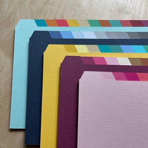 May include: A set of six colorful cardstock sheets with a textured finish. The sheets are arranged in a stack, with the top sheet being a light pink color. The other sheets are light blue, dark blue, yellow, burgundy, and a darker shade of pink.