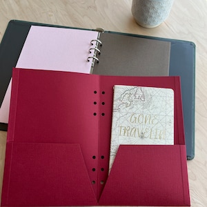 Può includere: Un raccoglitore rosso testurizzato a sei anelli con una pagina rosa e una pagina marrone. Il raccoglitore ha una tasca sul davanti con un piccolo quaderno all'interno. Il quaderno ha una mappa sulla copertina con il testo "Gone Traveling" in lettere dorate.