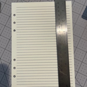 May include: A white sheet of lined paper with six evenly spaced holes down the left side. The paper is measured against a metal ruler with markings in inches and centimeters. The ruler is labeled "Staedtler Mars 963 53-12".