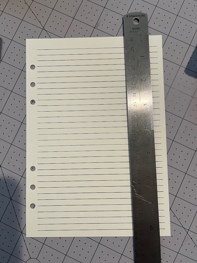 May include: A white sheet of lined paper with six evenly spaced holes down the left side. The paper is measured against a metal ruler with markings in inches and centimeters. The ruler is labeled "Staedtler Mars 963 53-12".
