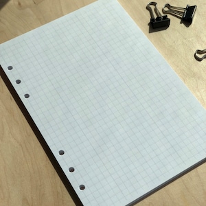 May include: A white gridded notebook with six holes on the left side of the page. The notebook is lying flat on a light brown wooden surface. Two black binder clips are on the surface to the right of the notebook.