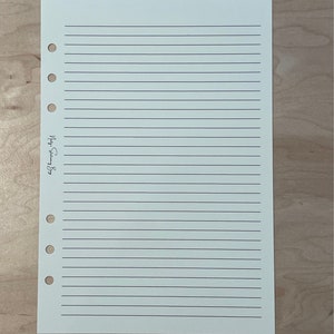 May include: A blank, lined, white paper sheet with a hole punch on the left side. The paper is for a planner or journal.