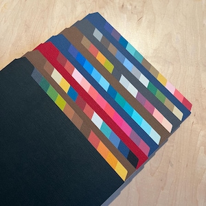 May include: A stack of colorful paper swatches, each with a different color gradient. The swatches are arranged in a fan shape, with the darkest colors at the bottom and the lightest colors at the top. The swatches are on a light wood surface.