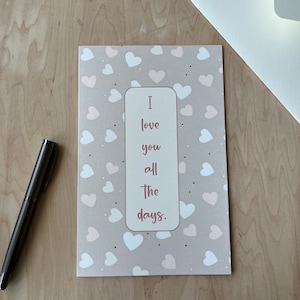 May include: A greeting card with a light gray background covered in white and pink hearts. The card has a rectangular cutout with the words "I love you all the days." A silver pen and white envelope are also present.