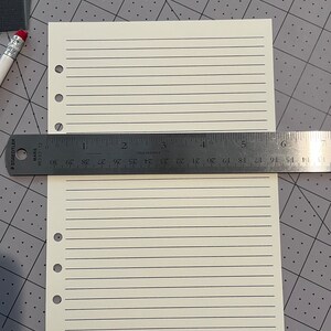 May include: A blank, off-white, lined paper sheet with six holes on the left side. The paper is lying on a gray gridded surface with a ruler measuring 7 inches.