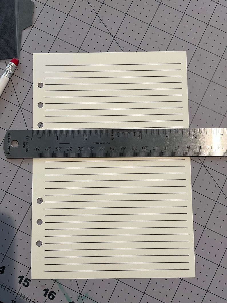 May include: A blank, off-white, lined paper sheet with six holes on the left side. The paper is lying on a gray gridded surface with a ruler measuring 7 inches.