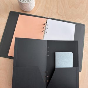 May include: A black ring binder with an open interior featuring peach and white pages, positioned behind a black two-pocket folder containing a small light blue notebook with a white floral pattern on its cover, and a small potted succulent plant.
