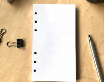 May include: A white, blank, dotted notebook with a black binding and six holes on the left side. The notebook is lying flat on a light wood surface.