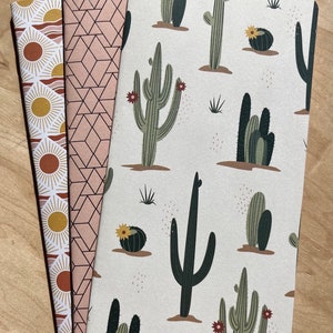 May include: Three notebooks with different patterned covers. The first cover has a repeating pattern of suns and waves in shades of orange, yellow, and brown. The second cover has a repeating geometric pattern in shades of pink and white. The third cover has a repeating pattern of cacti and flowers in shades of green, brown, and white.