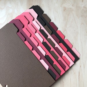 May include: A set of 12 colorful divider tabs in shades of pink, red, and brown. The tabs are made of textured paper and have a scalloped edge. The tabs are arranged in a fan shape, with the brown tab at the bottom and the pink tab at the top.