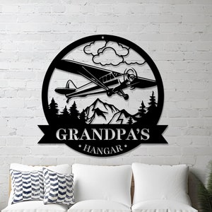 May include: Black metal sign with a silhouette of a plane flying over mountains and trees. The sign reads "Grandpa's Hangar".