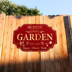 May include: Red metal garden sign with gold lettering that reads "Melissa's Garden Est. 2024 Veggies, Flowers, Herbs". The sign has a decorative scroll design and images of gardening tools and butterflies.