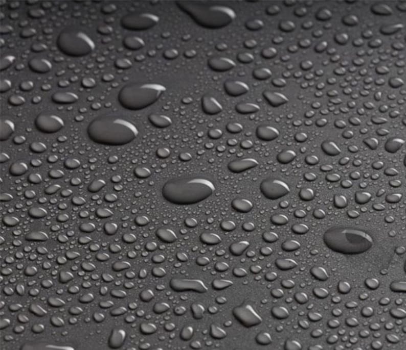 May include: Close-up of a dark gray surface covered in water droplets of varying sizes. The droplets reflect light, creating highlights and shadows. The surface appears smooth, with a subtle texture visible.