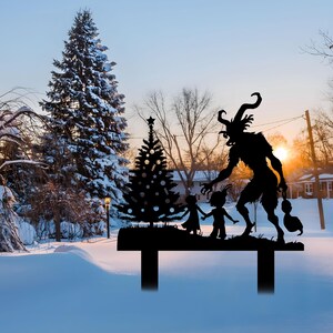 May include: A black metal yard stake featuring a silhouette of Krampus, a horned figure, with children and a Christmas tree. The stake is set in a snowy landscape with a sunset in the background.