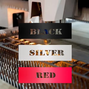 May include: Three rectangular metal signs with the words "BLACK", "SILVER", and "RED" cut out. The black sign is at the top, the silver sign is in the middle, and the red sign is at the bottom. The signs are in front of a metalworking machine.