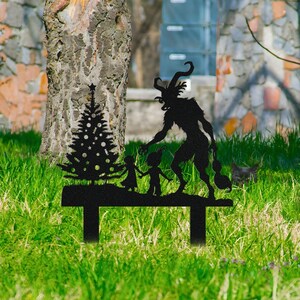 May include: Black metal yard art featuring a Christmas tree, two children, and a Krampus figure. The silhouette is set on a rectangular base with two legs, designed for outdoor display. The artwork is placed in a grassy yard.