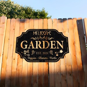 May include: Black garden sign with gold lettering that reads "Melissa's Garden Est. 2024 Veggies - Flowers - Herbs".