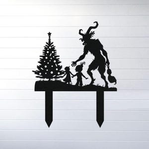 May include: Black metal Christmas yard stake featuring a silhouette of a decorated Christmas tree, two children, and a horned creature. The stake has two pointed prongs for easy placement in the ground. A festive holiday decoration.