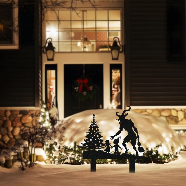 May include: Black metal yard art featuring a Christmas tree and a Krampus figure with children. The silhouette is set in a snowy outdoor scene, with a house and Christmas lights in the background. The yard art is designed to be placed in the ground.