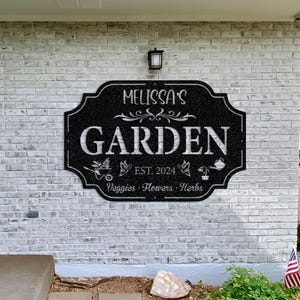 May include: Black metal garden sign with white text that reads "Melissa's Garden Est. 2024 Veggies - Flowers - Herbs".