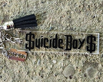 May include: A clear acrylic keychain with a black tassel and a black text overlay that reads "Suicide Boy$". The keychain is attached to a silver key ring.