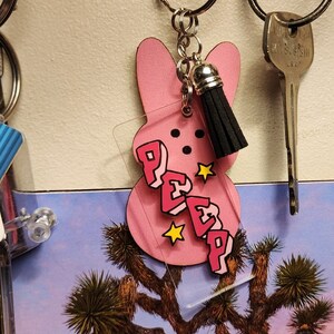 Puede incluir: Llavero rosa Peep con un borla negra. El llavero est&aacute; hecho de acr&iacute;lico y presenta un Peep rosa con la palabra "PEEP" escrita en letras rosas en la parte delantera. El llavero est&aacute; unido a un llavero plateado.