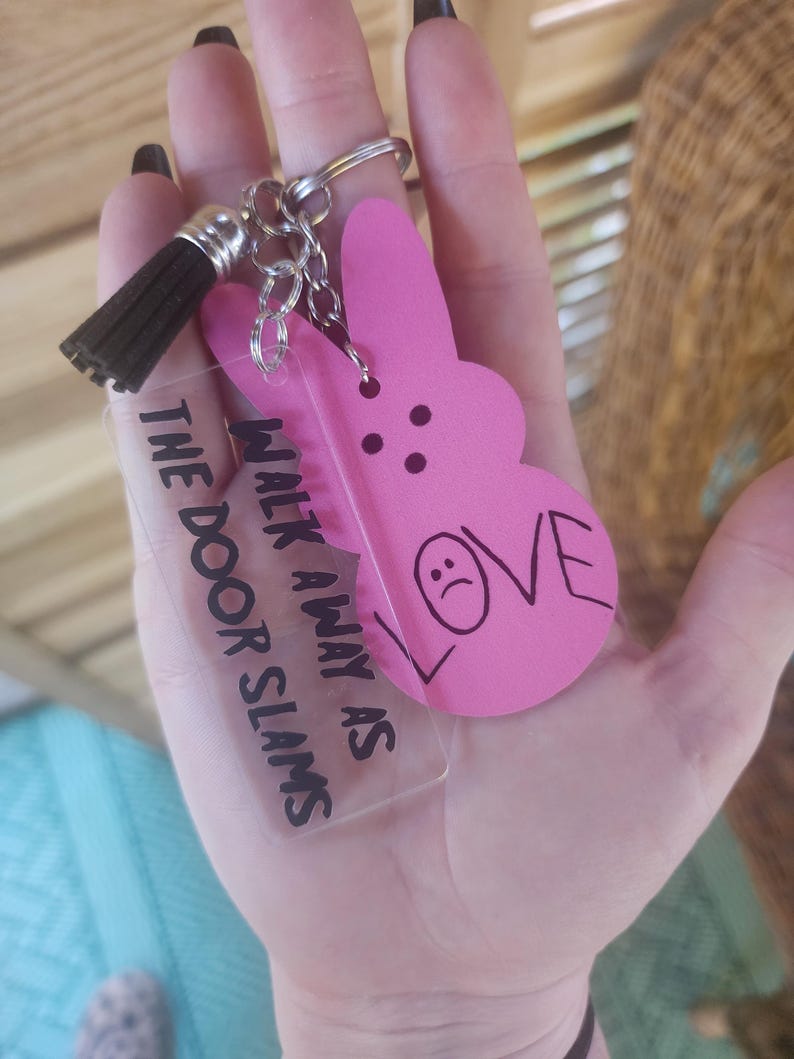 Op de afbeelding: Roze plastic konijn sleutelhanger met een zwarte kwast en een transparante acryl hanger met de tekst "Walk away as the door slams".