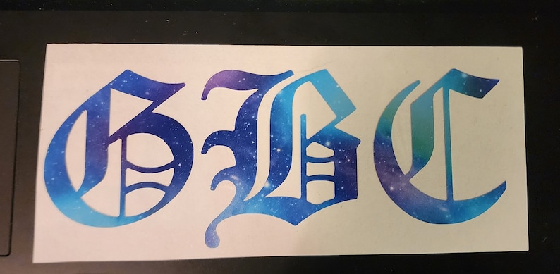 May include: A white sticker featuring the letters "GBC" in a gothic font. The letters are filled with a blue and purple galaxy design, creating a celestial effect. The sticker is rectangular.