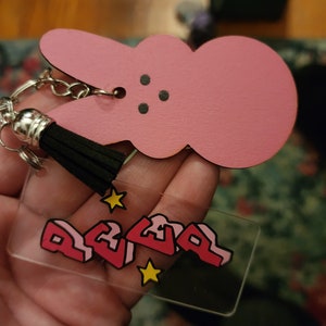 Puede incluir: Llavero de conejito rosa con borla negra y un dije de acr&iacute;lico transparente con la palabra "REVEL" en rosa y estrellas amarillas.