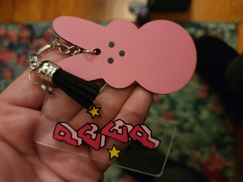 Puede incluir: Llavero de conejito rosa con borla negra y un dije de acr&iacute;lico transparente con la palabra "REVEL" en rosa y estrellas amarillas.