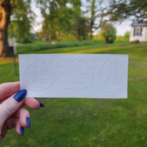 May include: A white sticker with the word "B!C" in a gothic font. The sticker is held up against a background of green grass and trees. The sticker is rectangular and has a grid pattern.