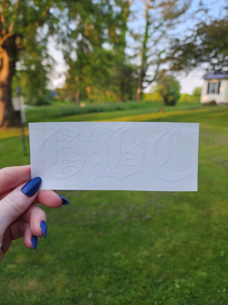 May include: A white sticker with the word "B!C" in a gothic font. The sticker is held up against a background of green grass and trees. The sticker is rectangular and has a grid pattern.