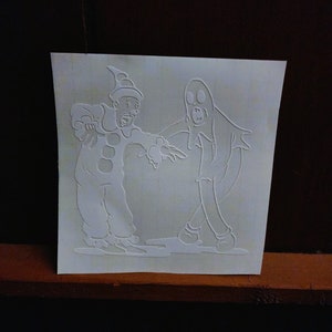May include: A white vinyl sticker featuring a cartoon ghost and a clown. The ghost is wearing a sheet and has a surprised expression. The clown is wearing a hat and a costume with a large red circle on the front. The sticker is perfect for adding a spooky touch to any Halloween decor.