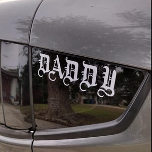 Puede incluir: Una calcoman&iacute;a de vinilo blanco con la palabra "DADDY" en fuente g&oacute;tica est&aacute; adherida a la ventana trasera de un veh&iacute;culo gris. La calcoman&iacute;a est&aacute; centrada y es claramente visible contra el cristal oscuro, con un &aacute;rbol.