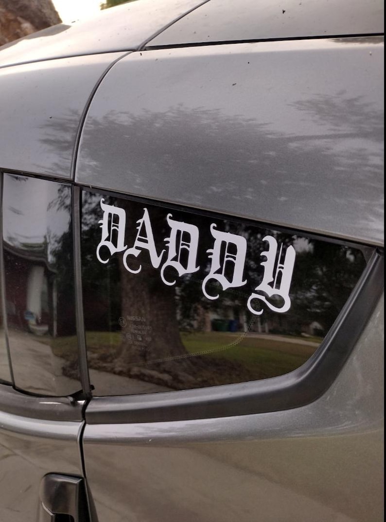 Puede incluir: Una calcoman&iacute;a de vinilo blanco con la palabra "DADDY" en fuente g&oacute;tica est&aacute; adherida a la ventana trasera de un veh&iacute;culo gris. La calcoman&iacute;a est&aacute; centrada y es claramente visible contra el cristal oscuro, con un &aacute;rbol.